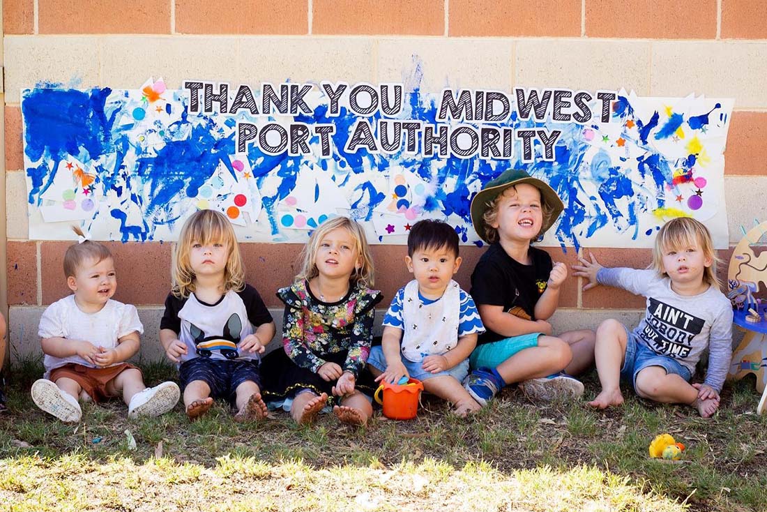 Six young children sitting in front of painted mural which says, 'Thank you Mid West Port Authority.'