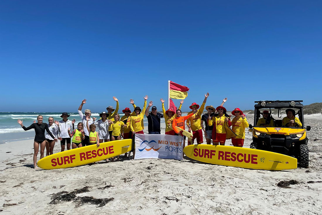Large group of people on beach standing with surf rescue boards, vehicle and Mid West Ports banner.