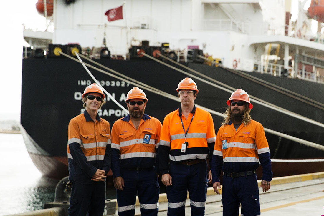 Four staff members standing on the berth looking at camera - black, white and red ship in background