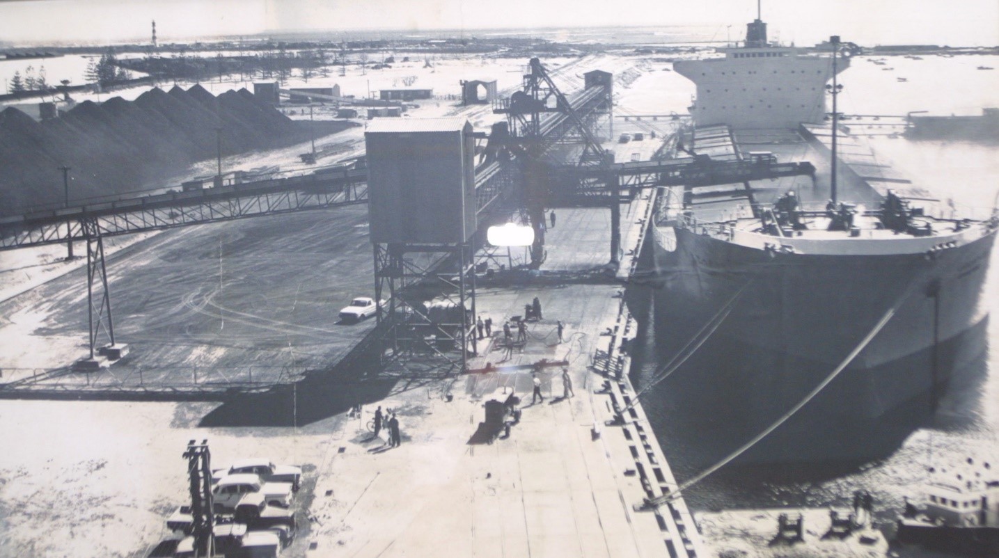 Black and white image of cargo ship on right with equipment on left loading mineral into ship. Piles of mineral appear on the left side.