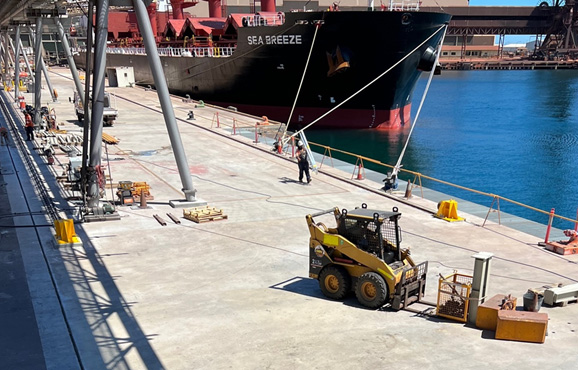 Concrete area of wharf with forklift operating in foreground, personnel walking and ship berthed in background.