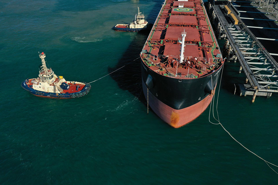 Front half of cargo ship with two tugboats alongside it.