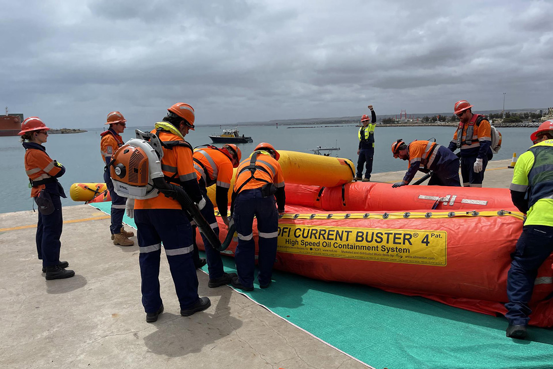 Eight people standing around red-orange, long, inflatable which says 'High Speed Oil Containment System' on yellow sign