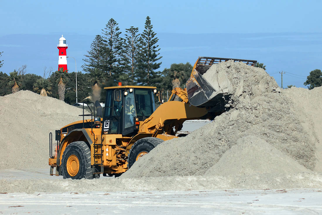 Front end loader lifting load of sand among piles of sand. Lighthouse in background.