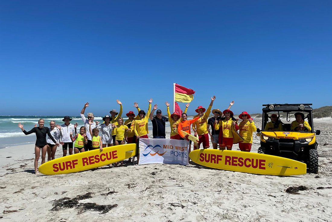 Group of adults and childrent waving while standing on the beach. Yellow surf rescue boards are in front with a yellow and red flag waving behind.