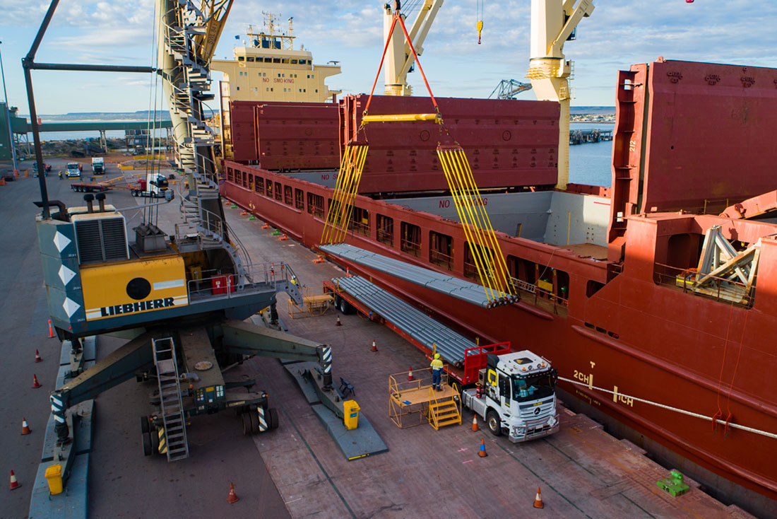 Pipes being lifted by yellow crane next to red cargo ship