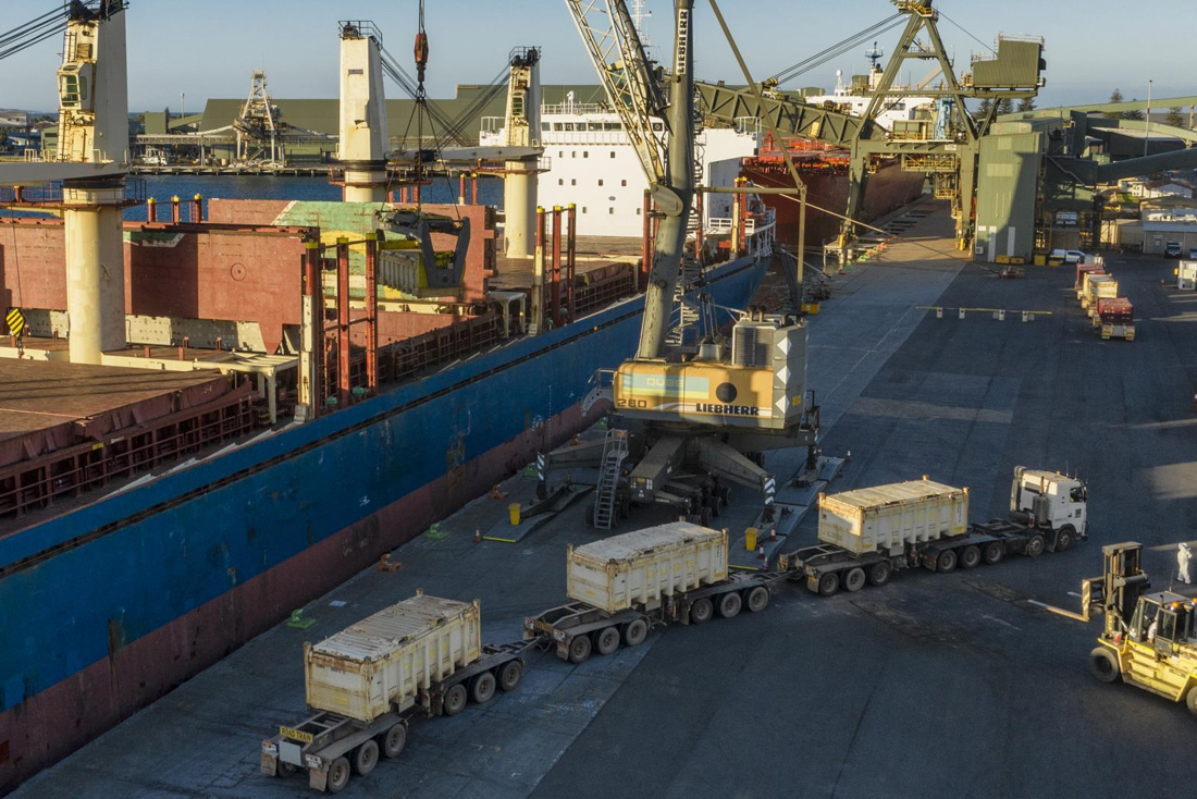 Truck curving in foreground around a forklift with crane and cargo ship behind it